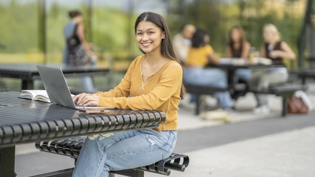 A young female University student, sits at a table outside as she studies between classes. She is dressed casually and smiling.